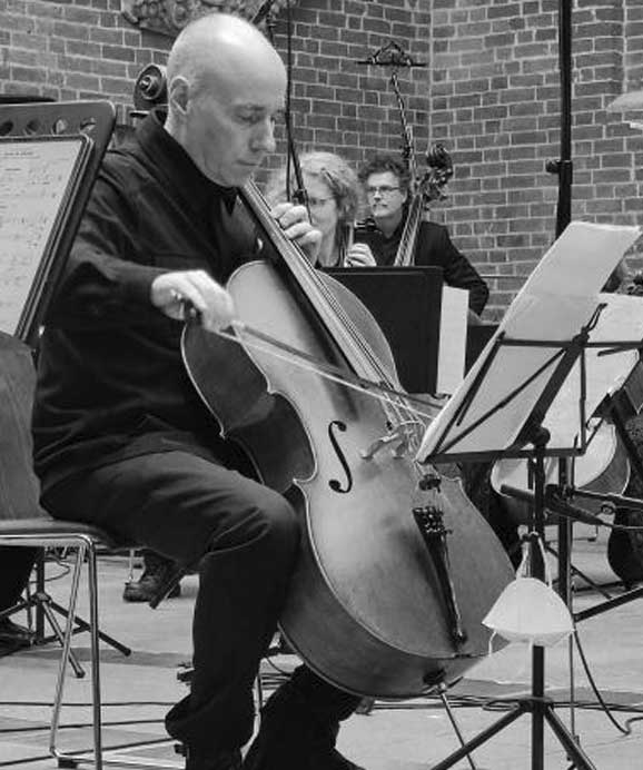 Schwarz-weiß-Foto eines Cellisten, der in einem Orchester in einem Gebäude mit Ziegelwänden spielt.