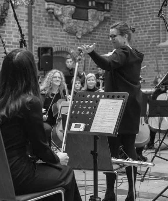 Dirigentin gibt Anweisungen an Orchester mit Streichern in historischem gemauertem Raum.