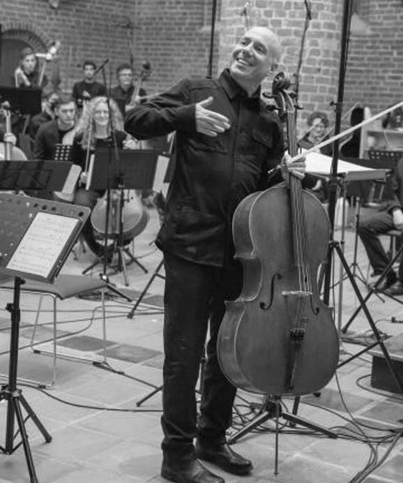 Ein lächelnder Cellist steht mit seinem Cello vor einem Orchester in einem Raum mit Backsteinwänden.