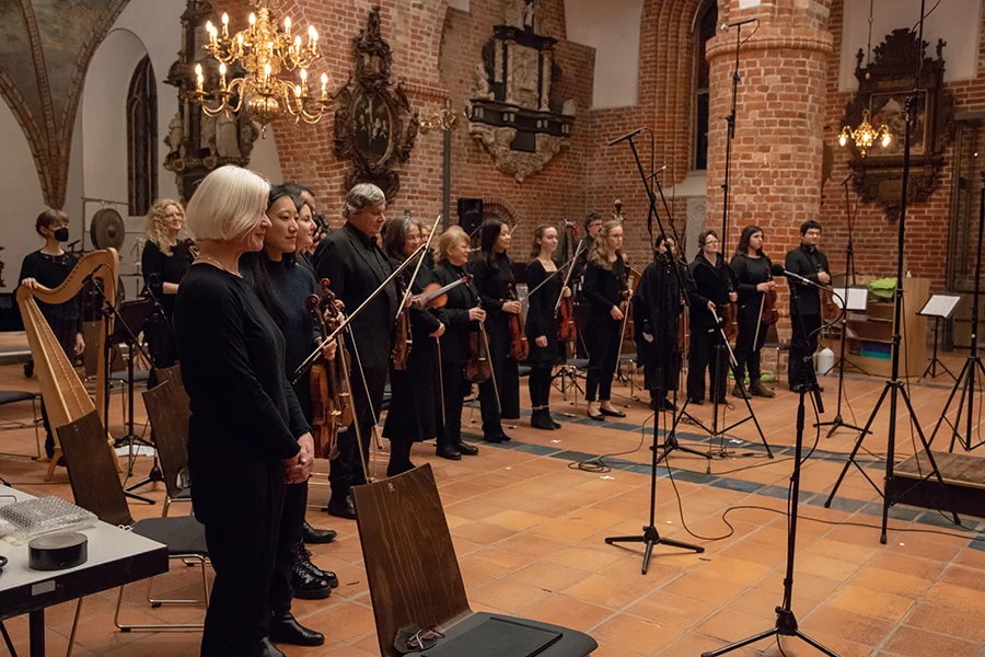 Orchestermitglieder in schwarzer Kleidung stehen mit ihren Streichinstrumenten in einer Kirche.