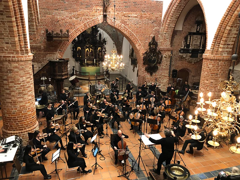 Ein Orchester spielt in einer Kirche mit roten Backsteinwänden und Kronleuchtern.