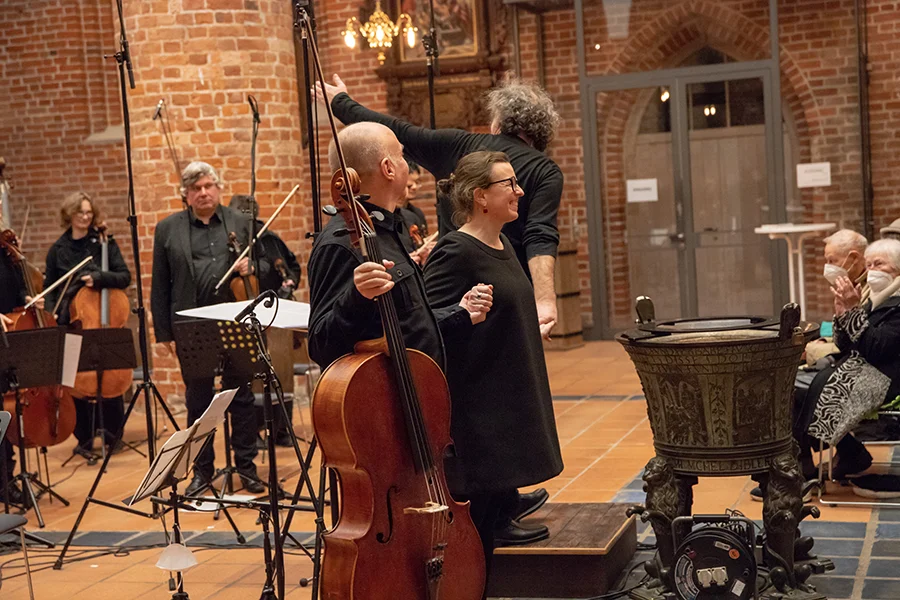 Ein Cellist und eine Dirigentin halten Händchen vor einem applaudierenden Publikum in einem Backsteingebäude.