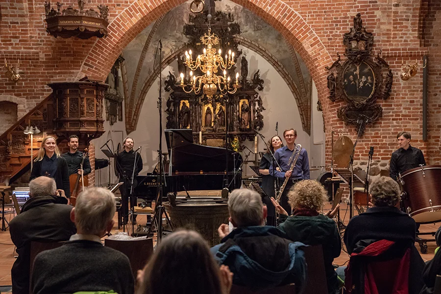 Musiker stehen vor Publikum in einer Kirche mit Backsteinwänden und Kronleuchter.