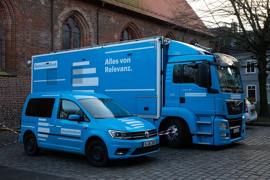 Zwei blaue Deutschlandfunk Fahrzeuge auf einem gepflasterten Platz vor Backsteingebäude.