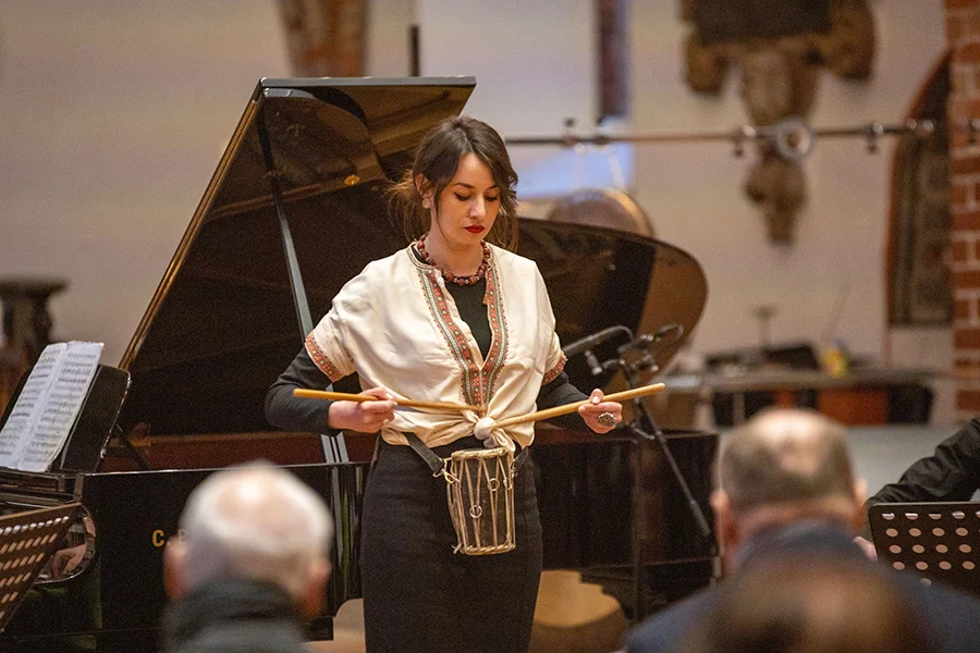 Frau spielt ein kleines traditionelles Trommelinstrument vor Publikum im Raum mit Klavier.
