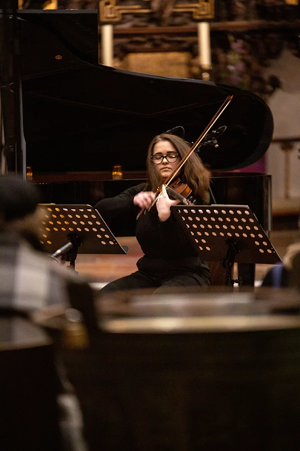 Frau spielt Violine vor Klavier bei einem Konzert in einem Raum mit Publikum.