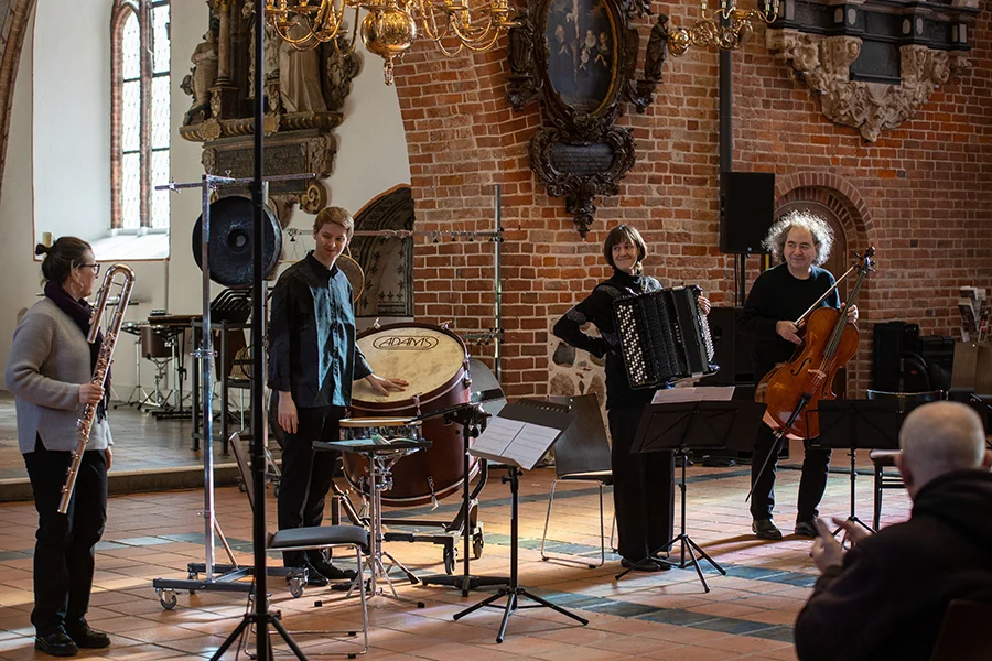 Vier Musiker mit Flöte, Schlagzeug, Akkordeon und Cello in einem Raum mit Backsteinwänden und Fenstern.