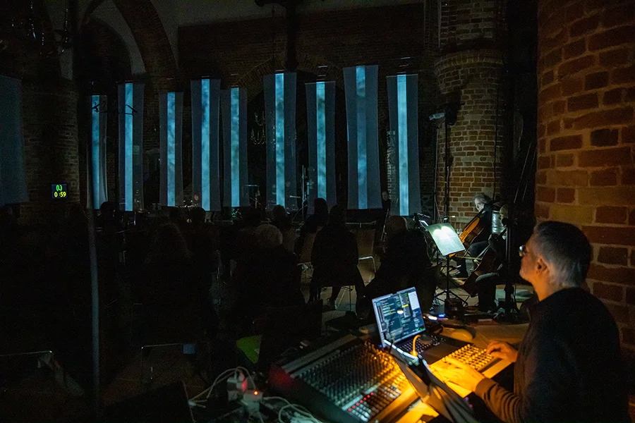 Techniker arbeitet an Audiomischpult bei einem Konzert in einem dunklen Veranstaltungsraum mit Musikern.