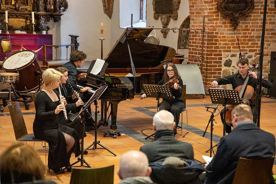 Ensemble mit Flöte, Klarinette, Klavier, Geige und Cello spielt in Konzertsaal vor Publikum.