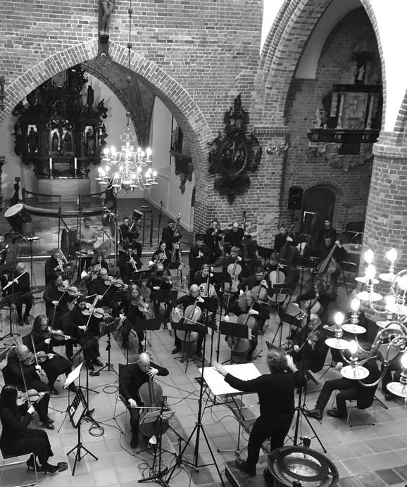 Schwarz-weiß-Foto eines Orchesters, das unter einem Kronleuchter in einer gotischen Kirche spielt.
