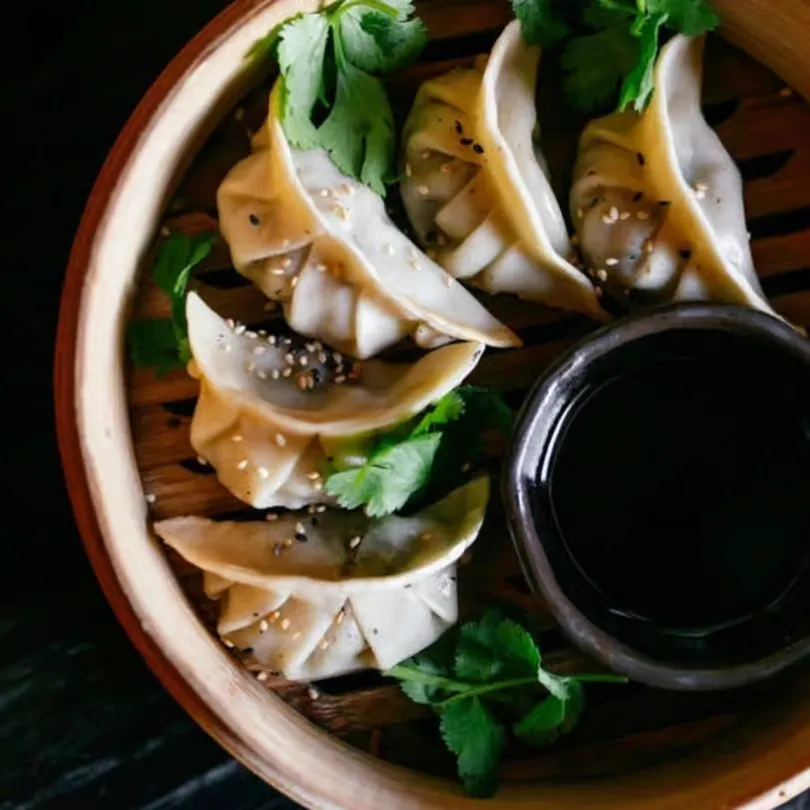 Steamed dumplings garnished with sesame seeds and cilantro in a bamboo basket with soy sauce.