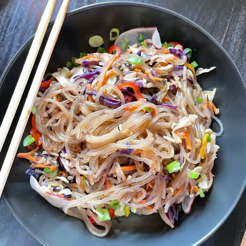 Black bowl of glass noodle salad with vegetables and chopsticks on a dark surface.