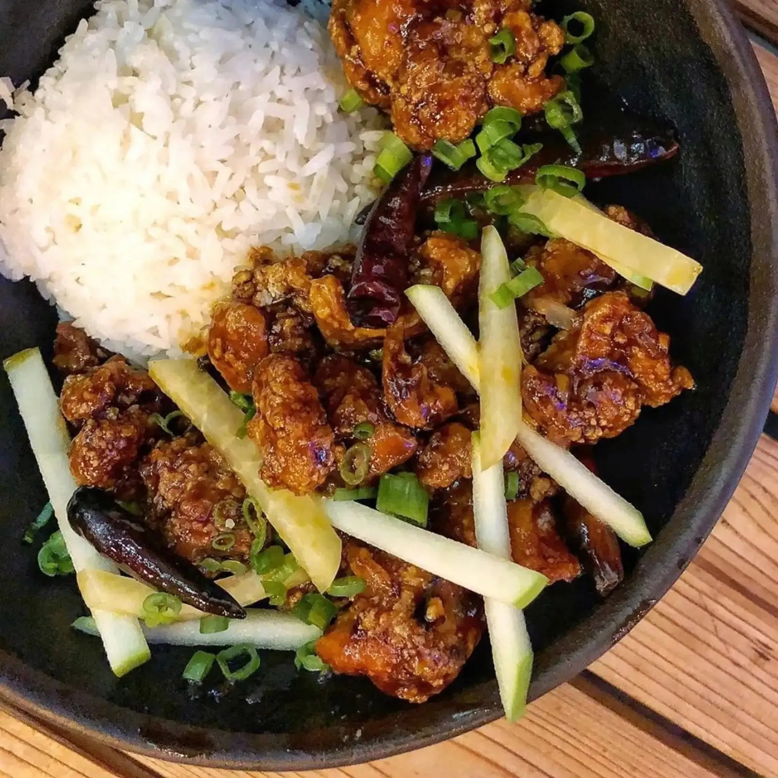 Black bowl with white rice, crispy fried chicken pieces, green onion, cucumber sticks, and dried chili peppers.
