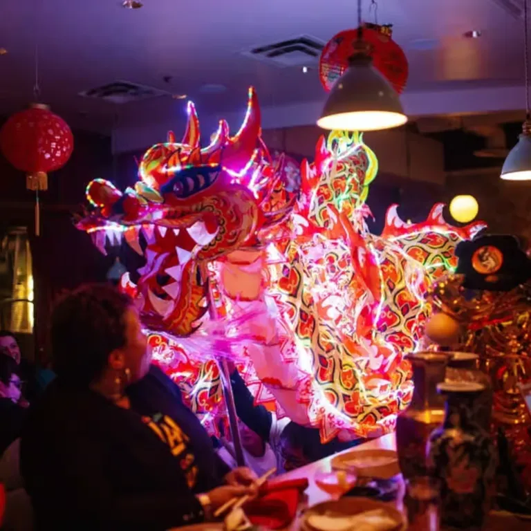 Performers holding a brightly lit, colorful dragon costume at an indoor event with red lanterns overhead.