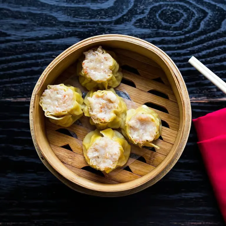Five steamed shumai dumplings in a round bamboo steamer basket on a dark wooden table.