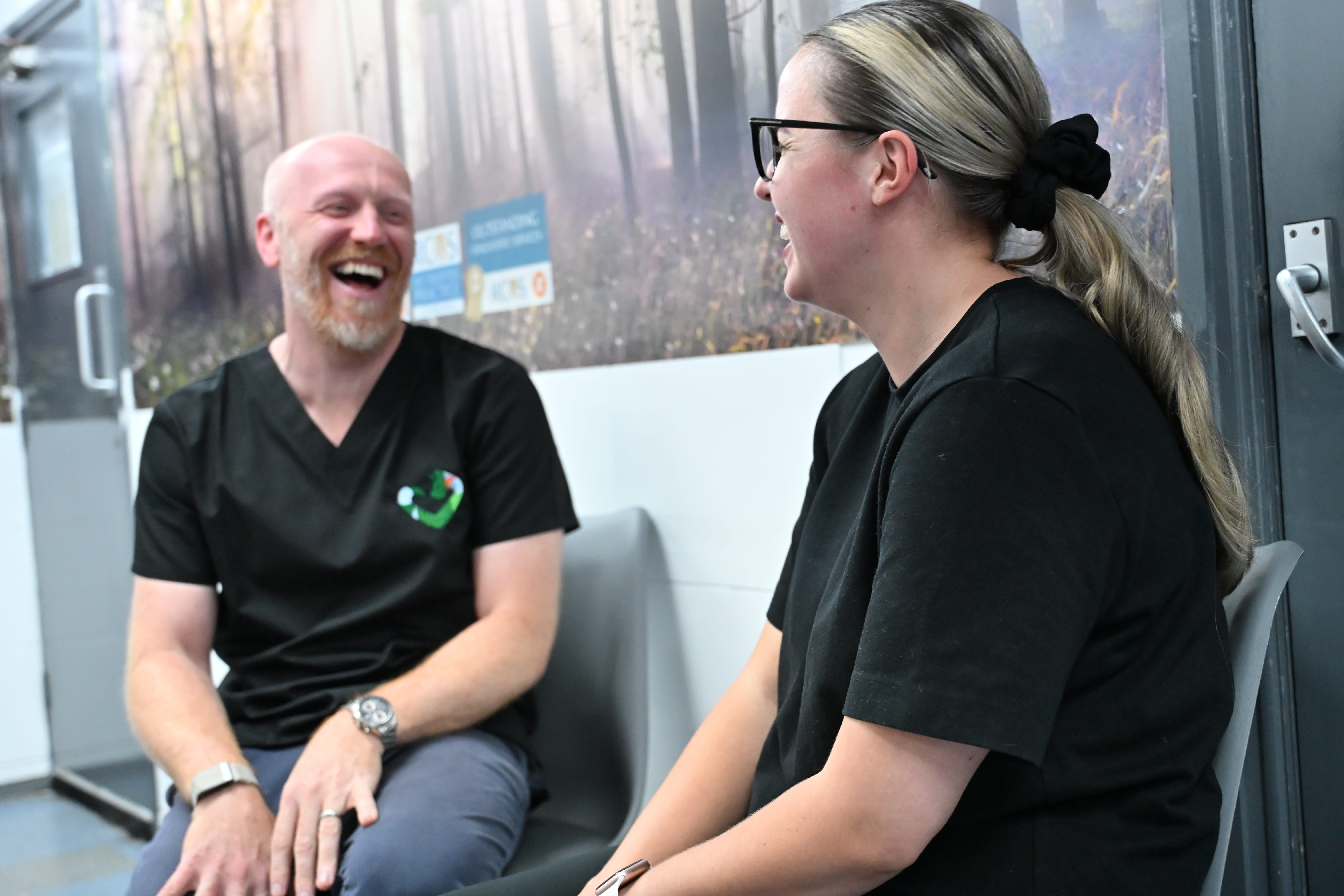 Two people in black shirts sitting on chairs, facing each other and laughing indoors.