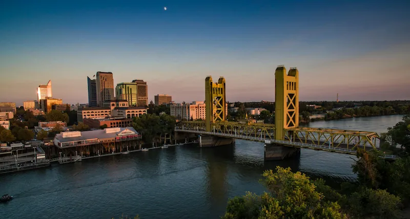 City skyline at dusk with a yellow vertical lift bridge over a river in the foreground.
