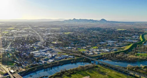 Aerial panoramic view of a city with rivers, roads, fields, and distant mountains under a clear sky.
