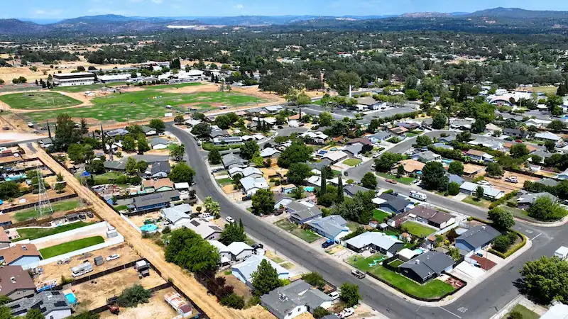 Aerial view of a residential neighborhood with houses, streets, trees, and distant hills under a blue sky.