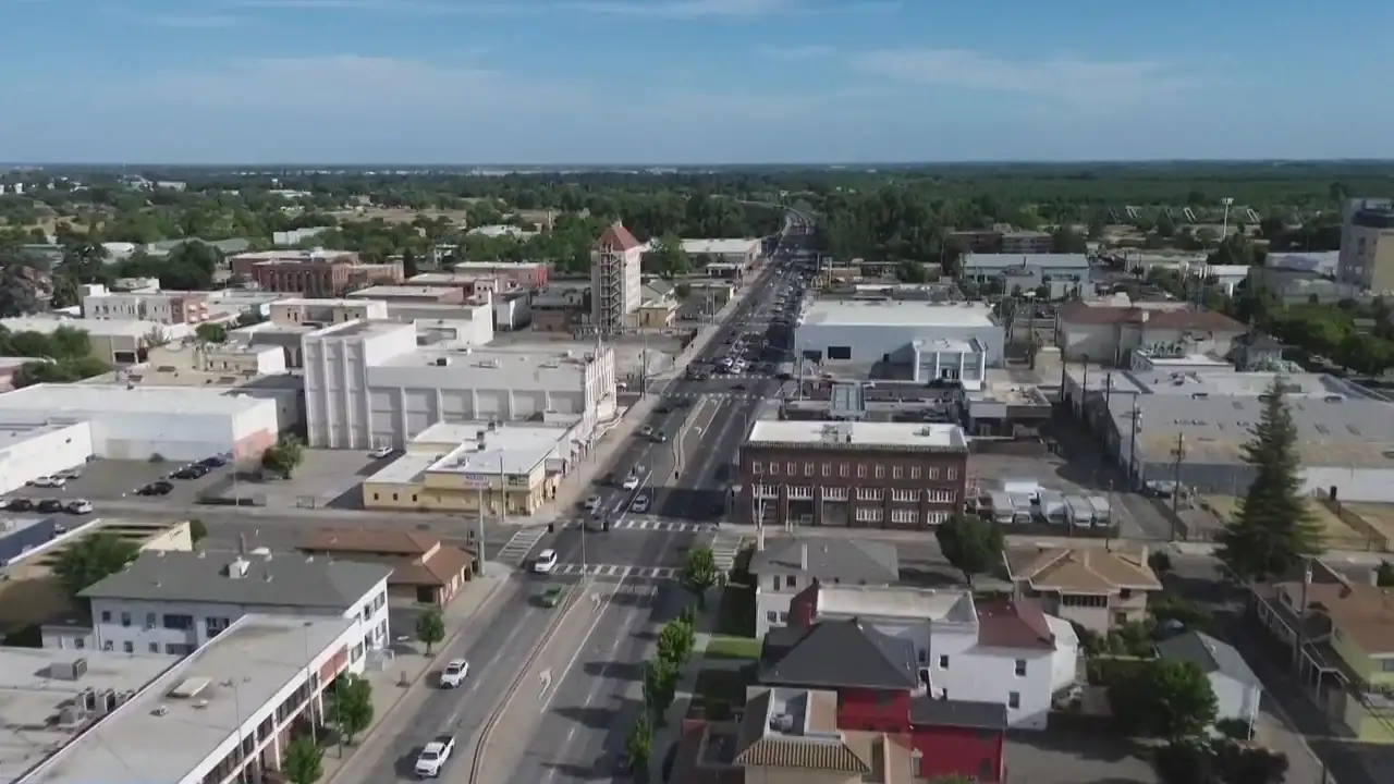 Aerial view of a small city with main road, various buildings, and green trees under a blue sky.