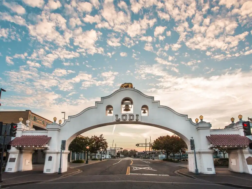 White archway with bells and 'LODI' sign over an empty street under a cloudy sky at sunset.