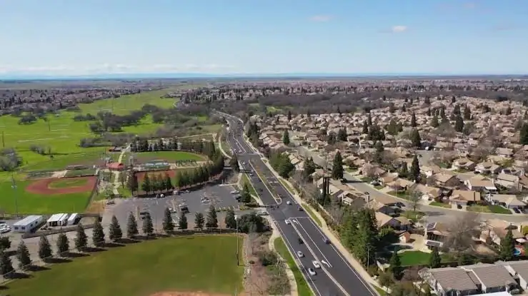Aerial view of a busy road with cars between a sports field and dense residential neighborhood.
