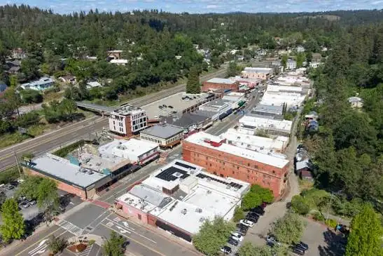 Aerial view of a small town with commercial buildings, roads, parking lots, and surrounding forested hills.