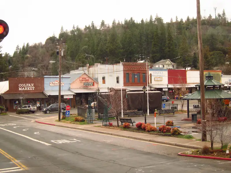 Small town street with shops including Colfax Market and Colfax Pharmacy, trees on hillside behind.