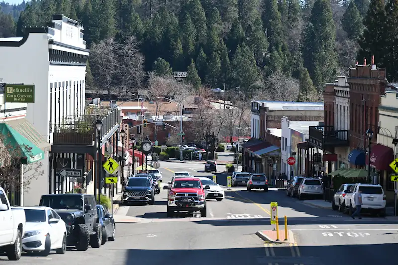 Main street in a small town with cars, shops, and tall evergreen trees in the background.