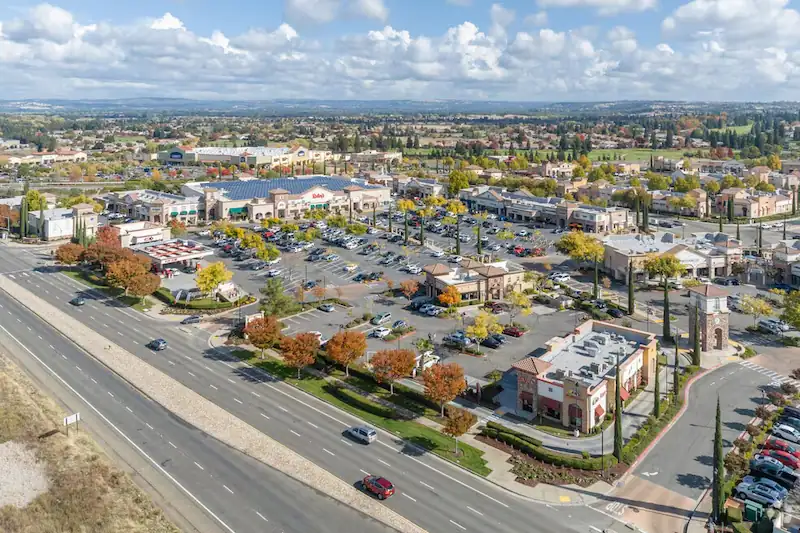 Aerial view of a commercial shopping center with parking lots and surrounding roads under a partly cloudy sky.