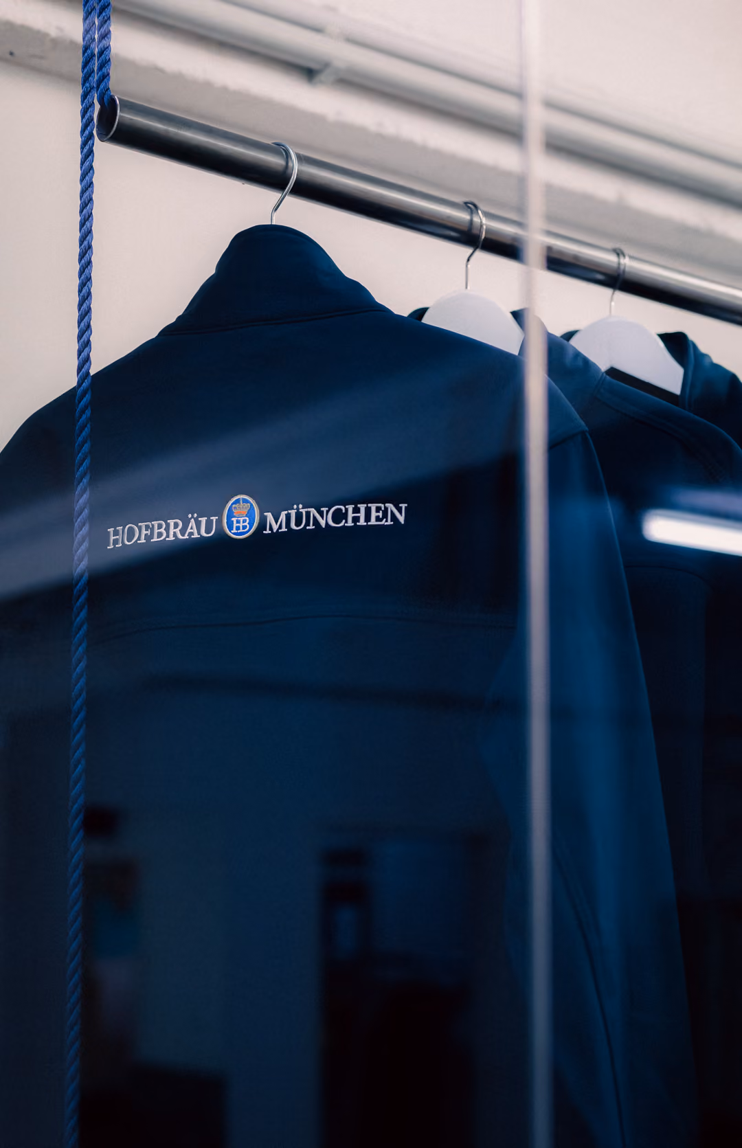Dark blue jacket with Hofbräu München logo hanging on a metal rack with hangers.
