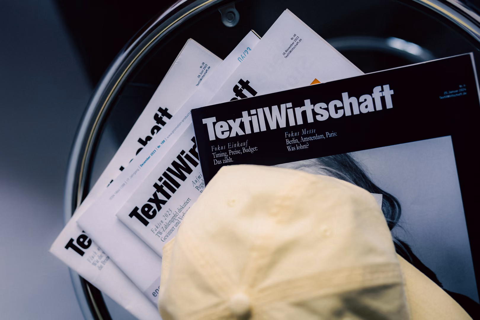 Stack of TextilWirtschaft magazines in a round metal tray with a light yellow cap on top.