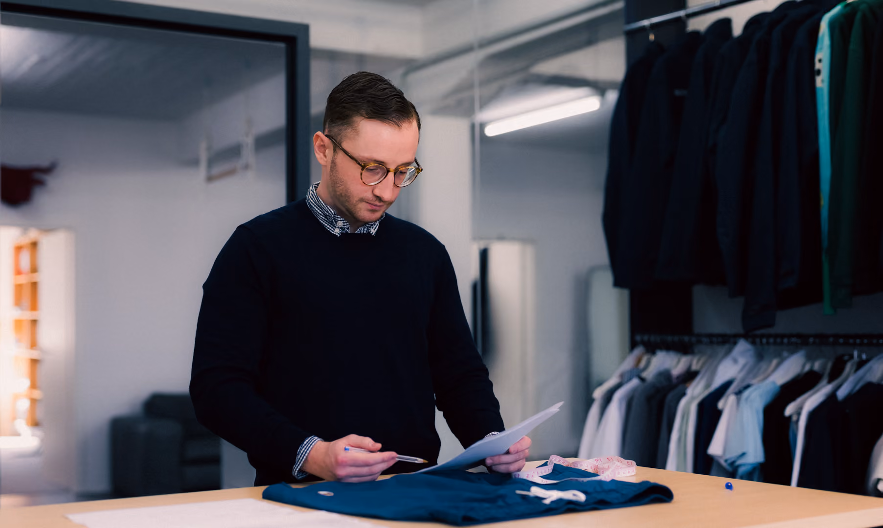 Man wearing glasses examines a document while standing at a table with clothes and measuring tape.