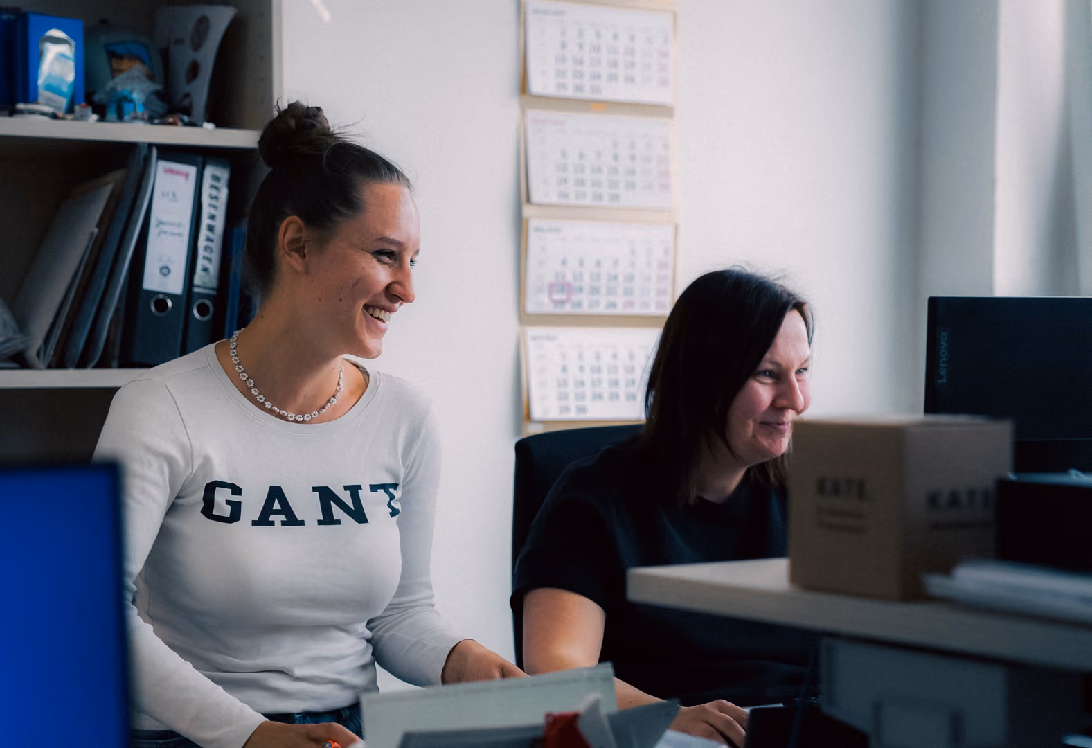 Two women smiling and working together at desks in an office setting with calendars on the wall.