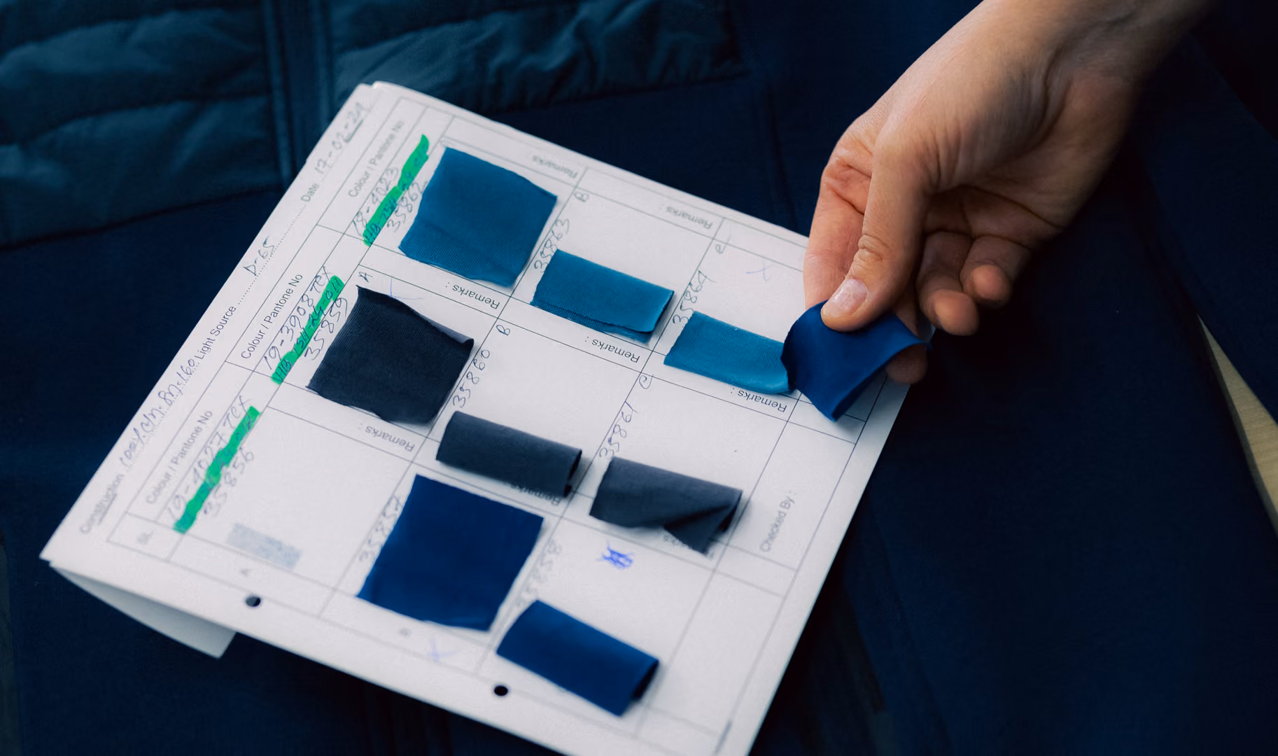 Hand holding a blue fabric sample above a paper with multiple blue fabric swatches and notes.