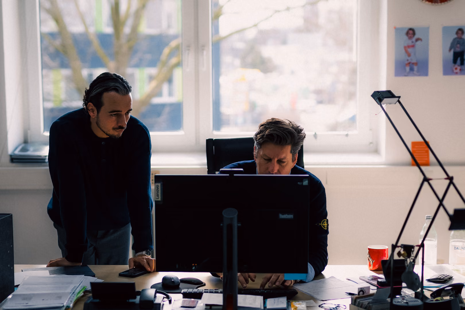 Two men working together at a desk with a computer in a bright office with large windows.