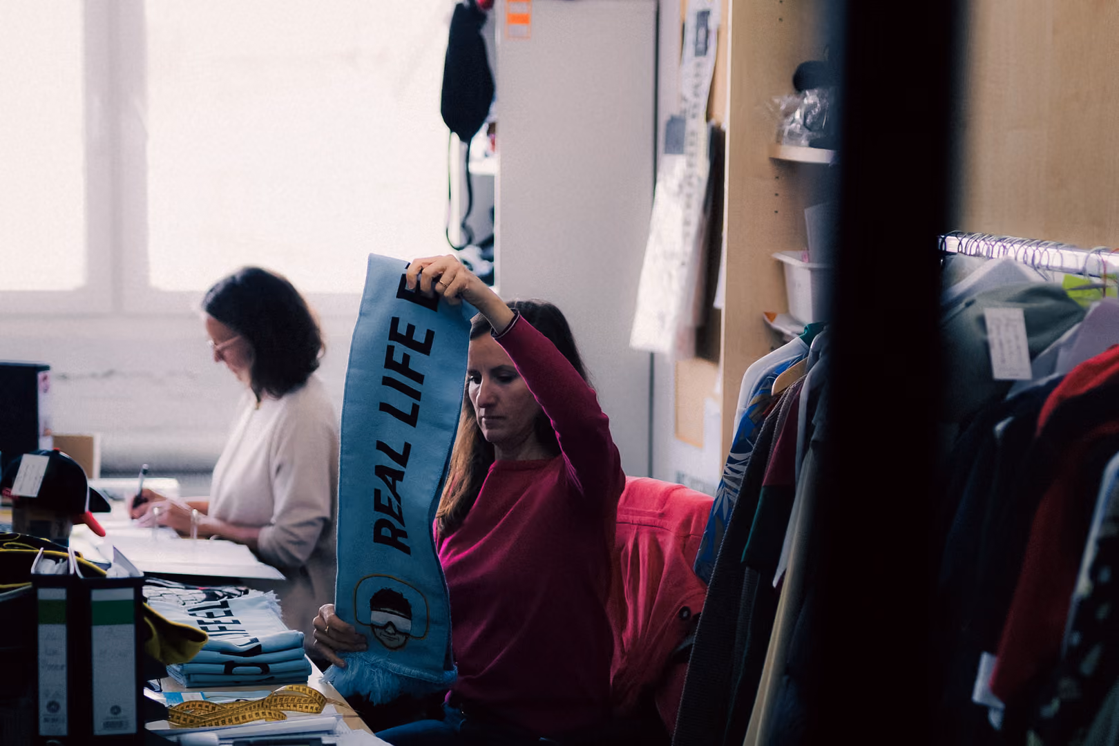 Woman in a pink sweater examining a blue scarf with 'REAL LIFE' text in a cluttered room.
