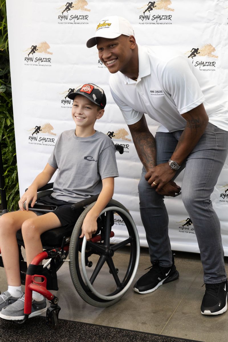 Smiling man in white shirt and cap poses beside smiling boy in wheelchair against Ryan Shazier backdrop.