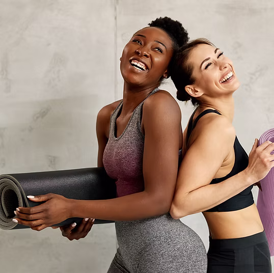 Two women in workout clothes smiling and holding Pilates mats while standing back to back.