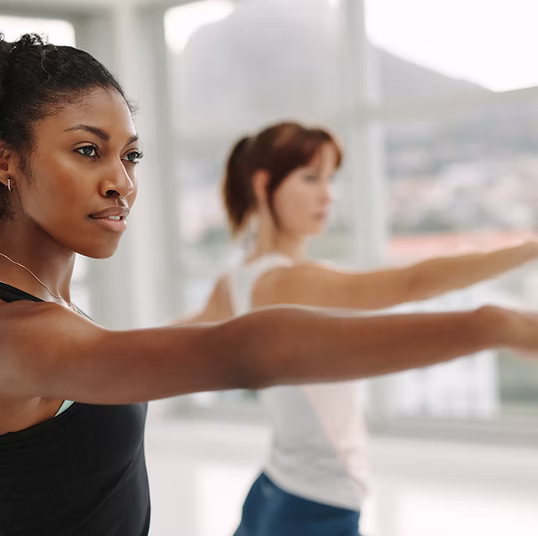 Two women practicing Warrior Two yoga pose in a bright room with large windows.