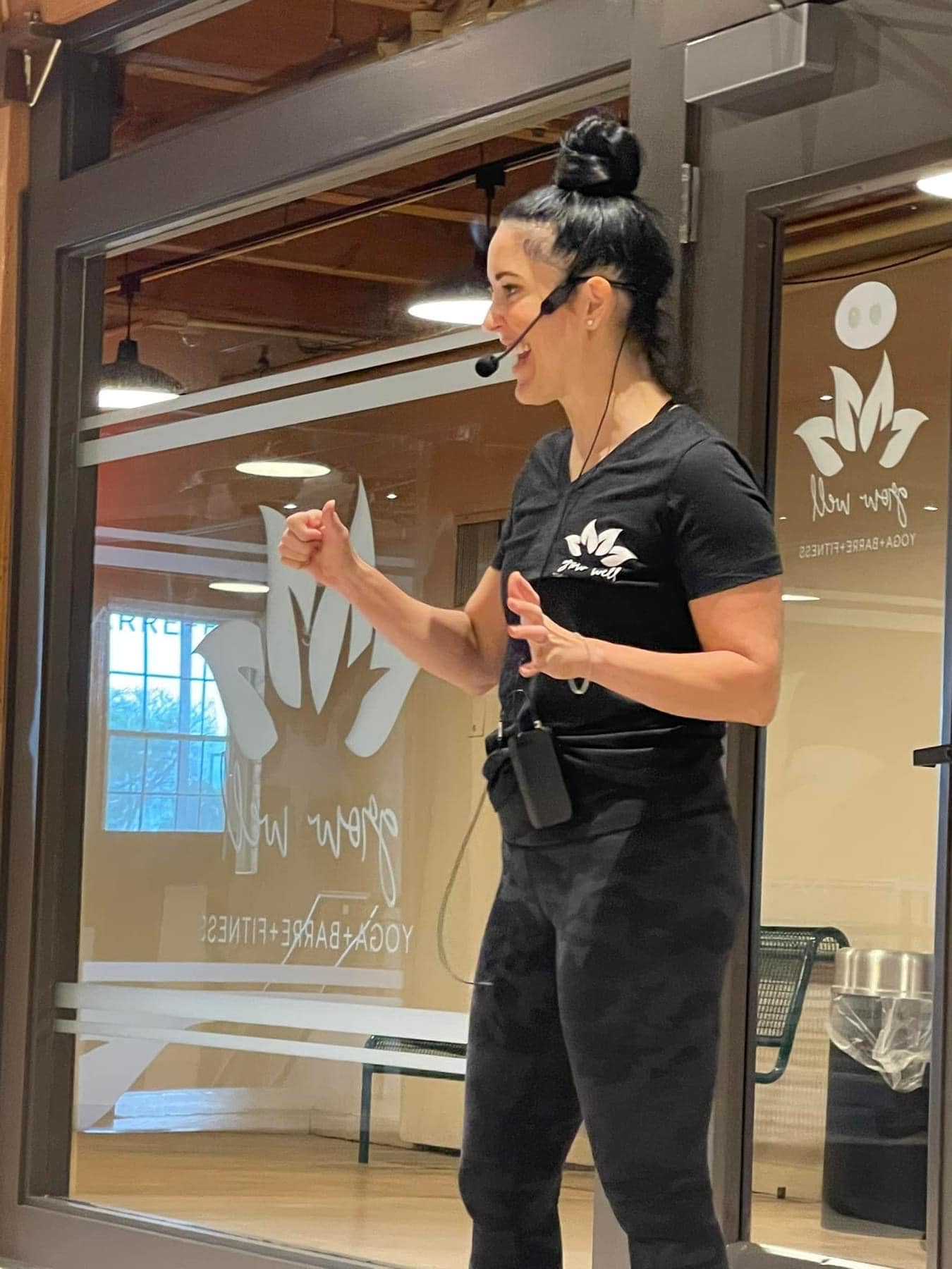 Woman fitness instructor with headset giving a thumbs up inside a yoga and fitness studio.