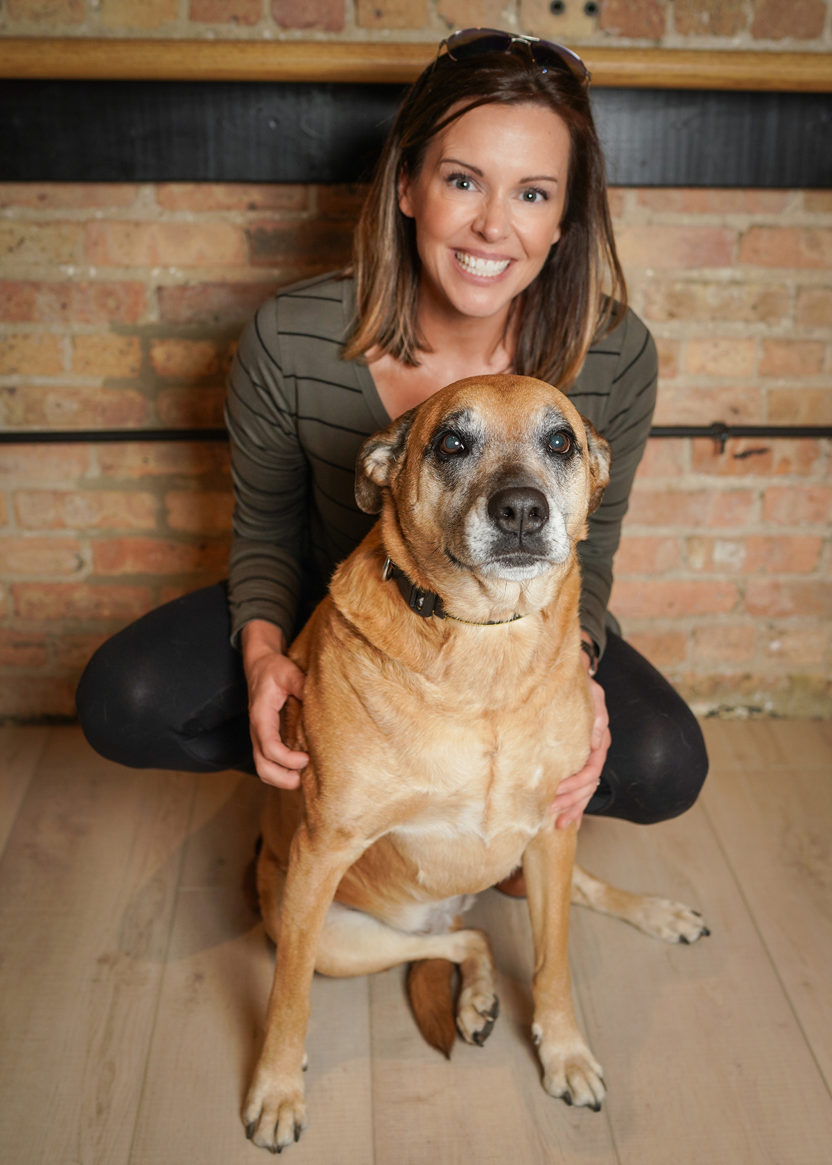 Smiling woman crouches behind large tan dog sitting on wooden floor with brick wall background.