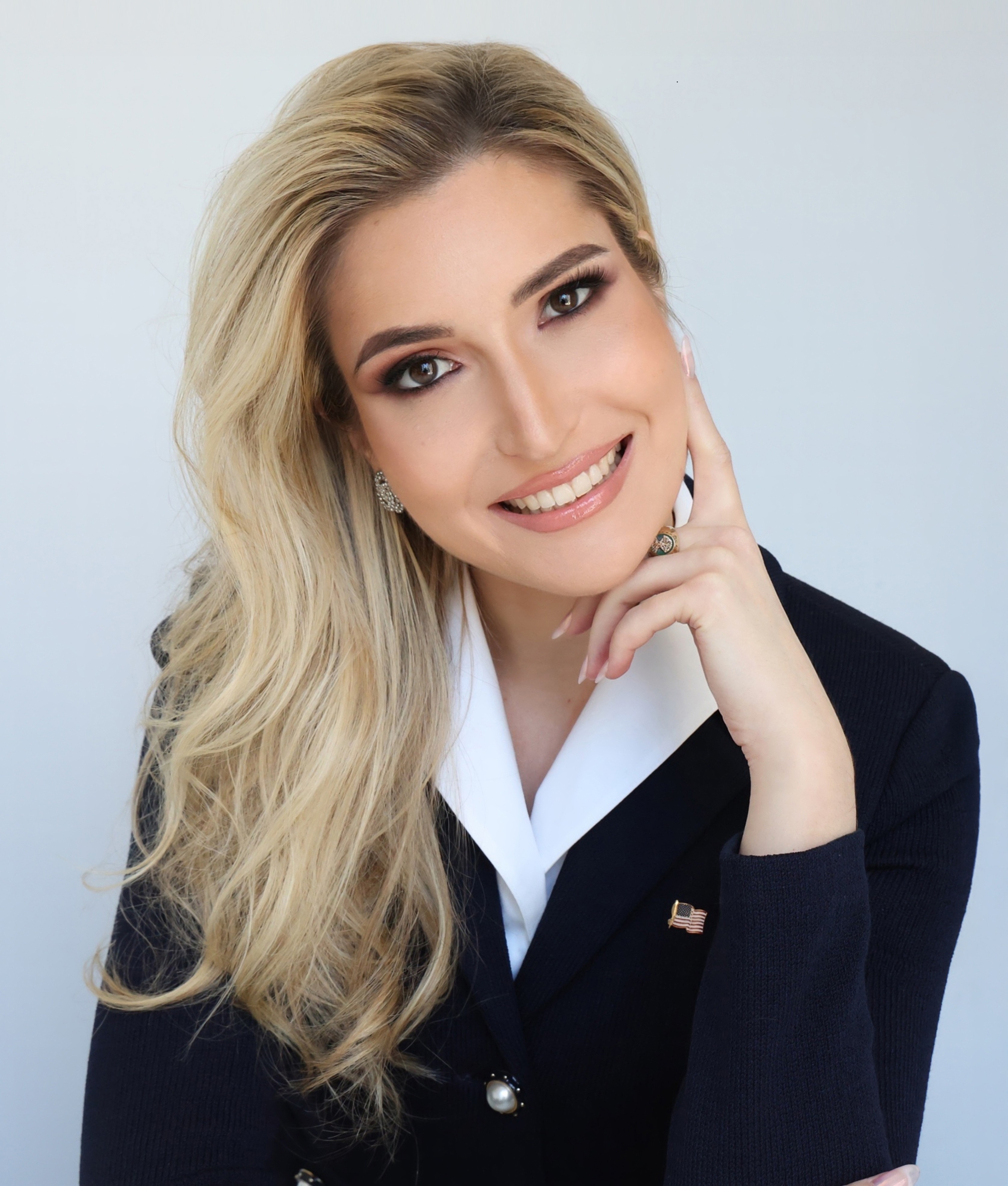 Smiling woman with blonde hair wearing a dark blazer with a US flag pin on a white background.