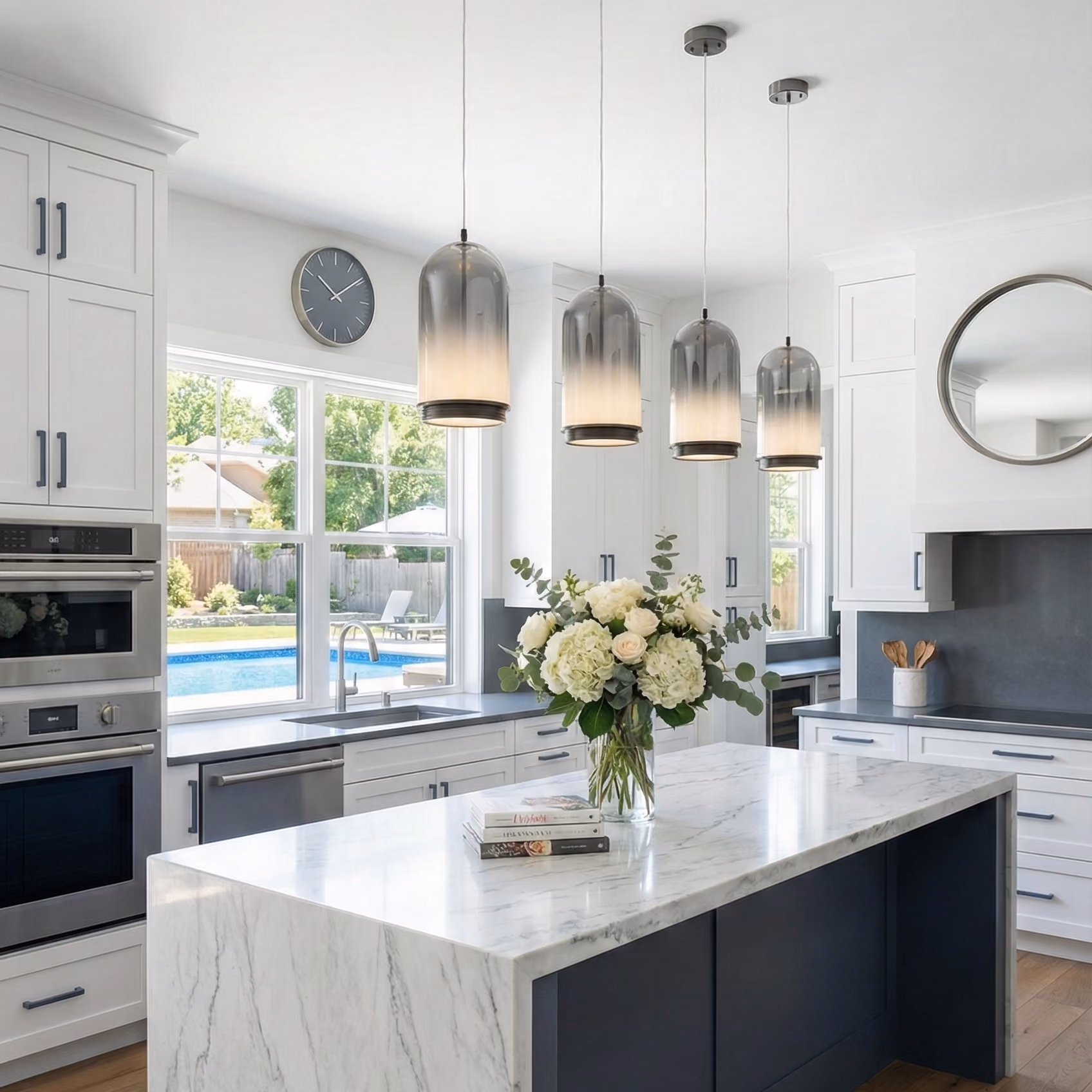 Modern kitchen with white cabinetry, a large marble island with a vase of white flowers, gray pendant lights hanging above, and a window showing a backyard pool.
