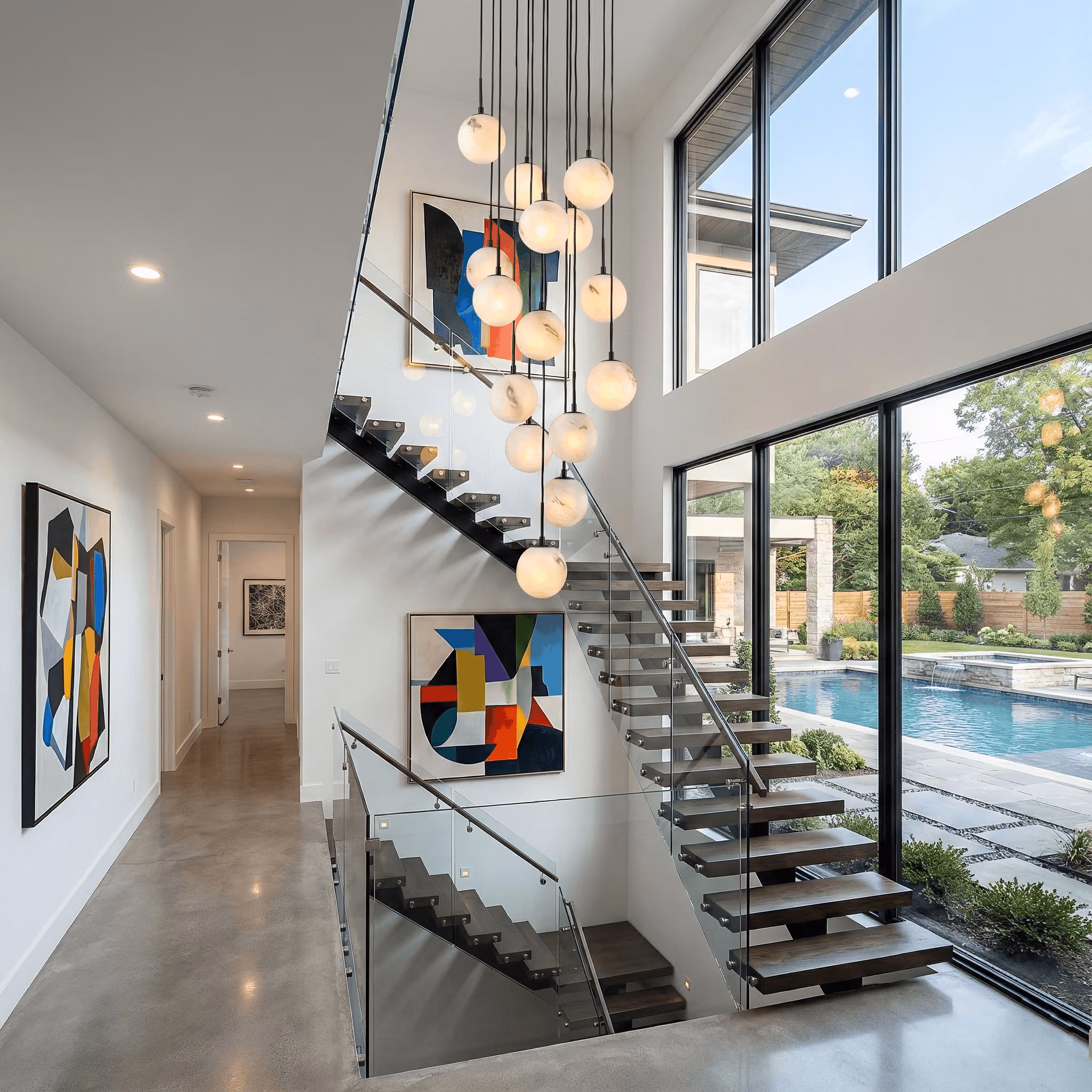 Modern interior with a floating wooden staircase featuring glass railings beside a large window overlooking a pool and garden, decorated with multiple hanging globe lights and abstract wall art.