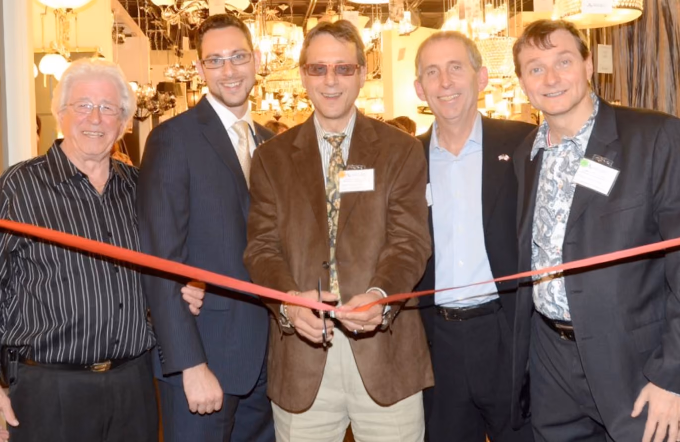 Five men standing indoors at a ribbon-cutting ceremony, with the man in the center cutting a red ribbon using scissors, surrounded by warm lighting fixtures.