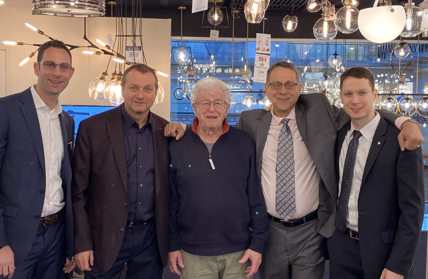 Five men standing closely together indoors in front of a large window and hanging decorative lights, smiling at the camera; four men wear suits and one wears a navy sweater with a red collar.