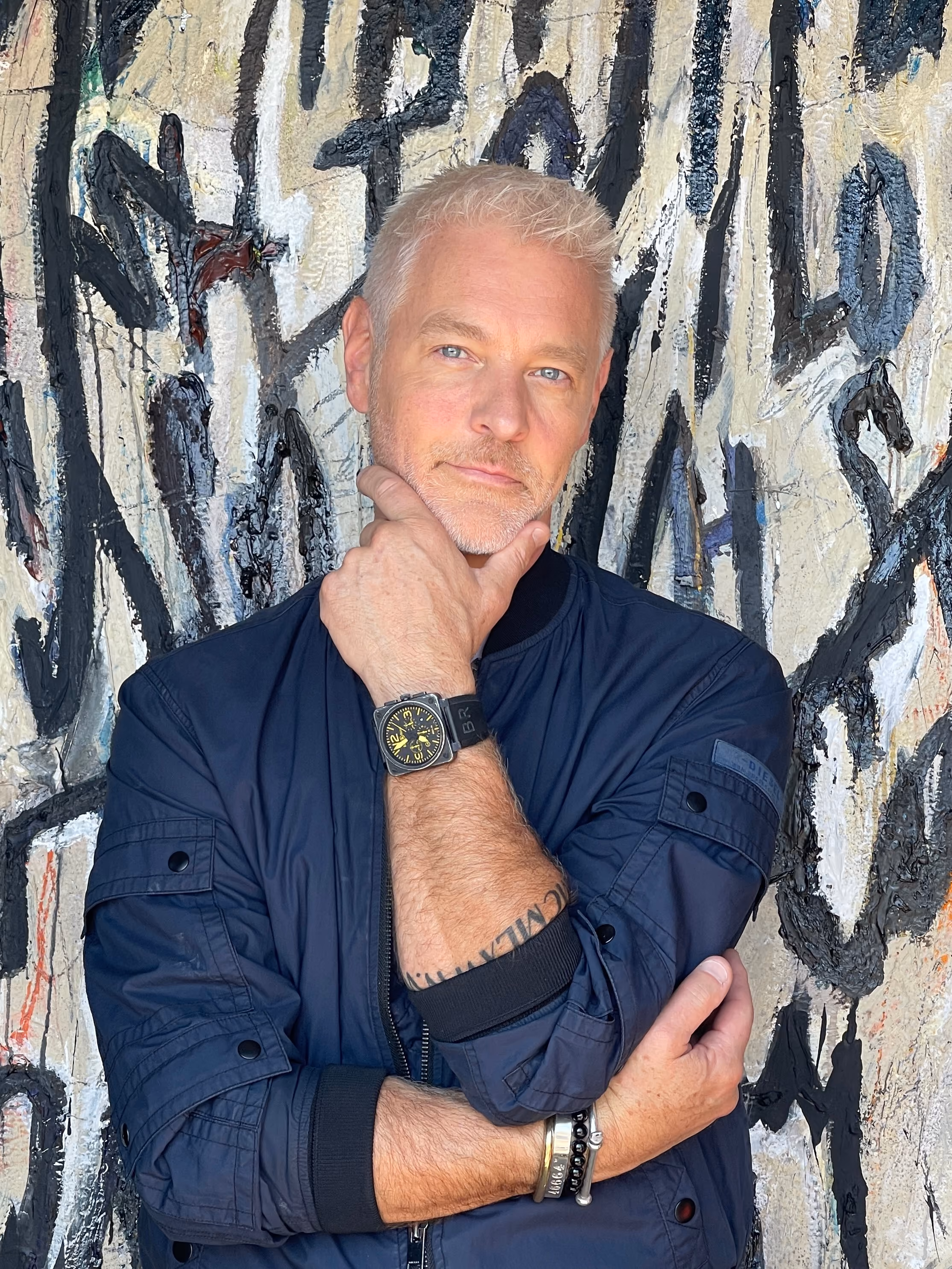 Man with short white hair and light beard wearing a dark blue jacket poses with one hand on his chin and the other crossed, standing against a graffiti-covered wall.