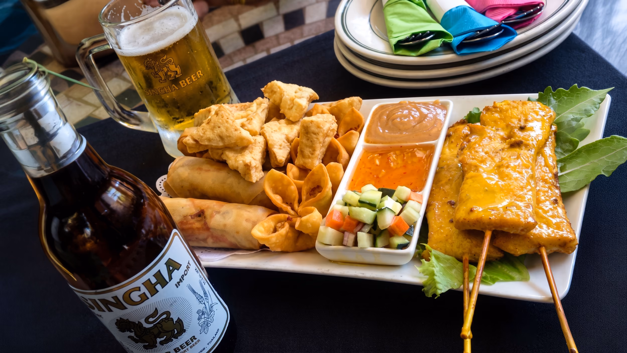 Plate of Asian appetizers with spring rolls, satay skewers, dipping sauces, and a glass of beer.