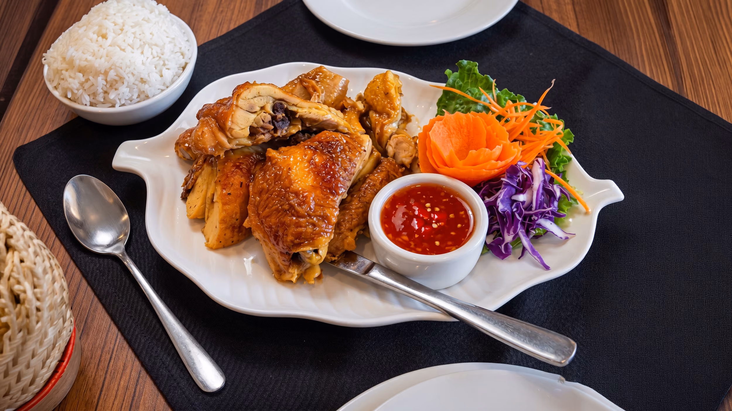 Plate of roasted chicken with chili sauce, shredded cabbage, carrot rose, lettuce, and a bowl of white rice.